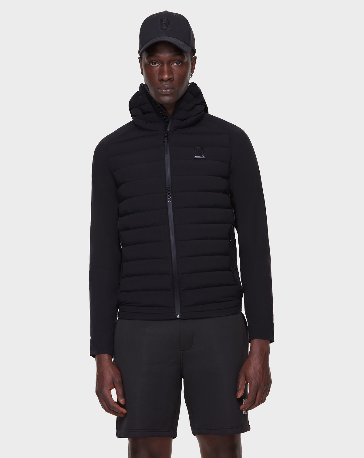 A person wears the RUDSAK JARRY - 6122007 BLACK men’s padded windbreaker jacket with a hood, paired with black shorts and a baseball cap, standing against a plain white background—a cold-weather staple.