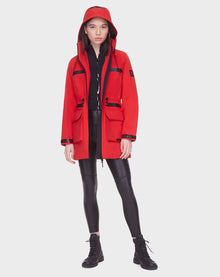 A woman faces forward in a Rudsak CAMY - 8122002 POPPY RED water-resistant coat with black zippers and accents, paired with black leggings and lace-up boots, set against a plain white background.